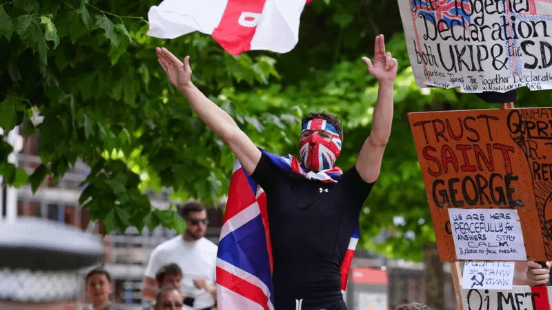 A man wearing a Union Jack mask, with a flag tied around his neck like a cape, doing a Nazi saute with his right hand and a finger gun with his other.